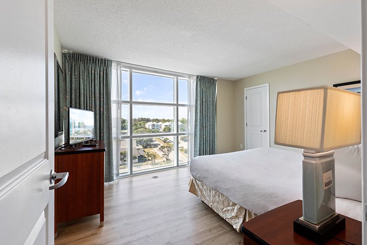 A hotel room with a large window, a bed, a TV on a dresser, and a lamp on a nightstand.