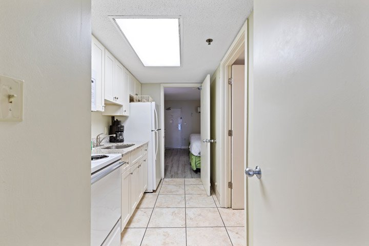 A small kitchenette with white appliances and tiled floor, an open doorway leading to a bedroom with a bed.