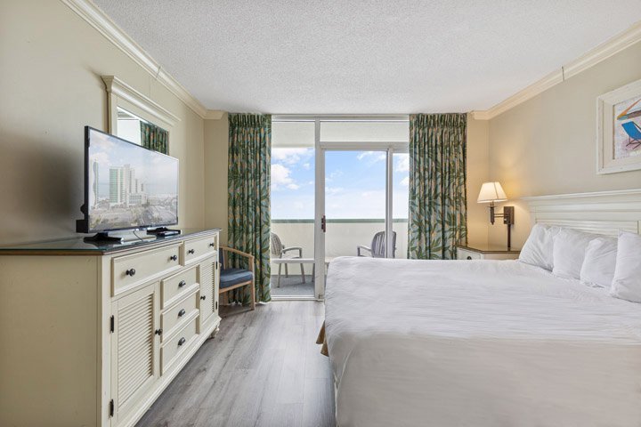 A hotel room with a large bed, TV on a dresser, and a balcony door with a view.