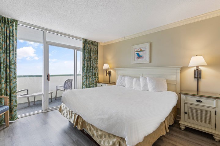 A hotel room with a large bed, two nightstands, and a sliding glass door leading to a balcony.